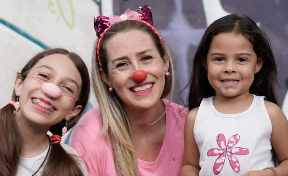 The image features a group of three people, consisting of two women and a little girl, who are all smiling and posing for a picture. One of the women is wearing a pink shirt, and the girl is holding a flower.