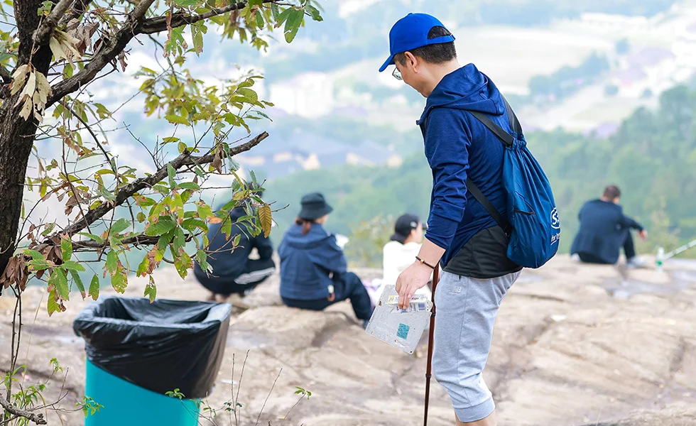 The image features a man standing on a mountain top, holding a stick and collecting waste.