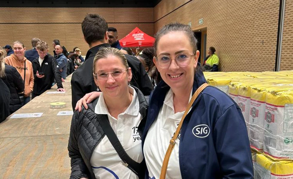 Two women smiling at a community event with stacked supplies in the background (photo)